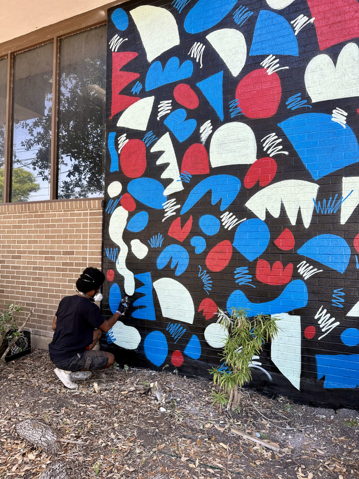 Kel Brown spray painting a bold red, blue and cream abstract mural on a brick wall